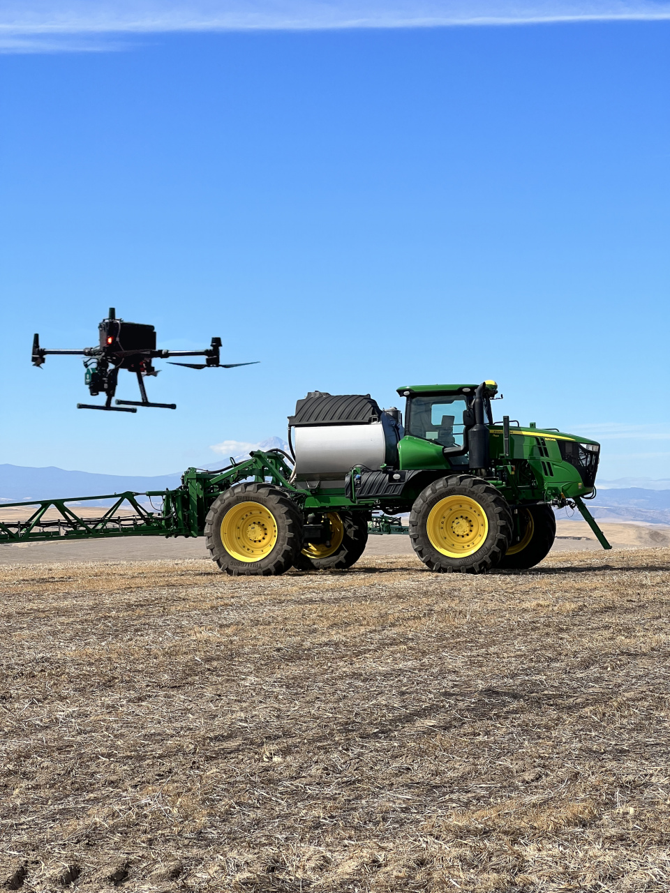 Agcopter drone with weed-detection sensor flying beside a John Deere sprayer in a stubble field